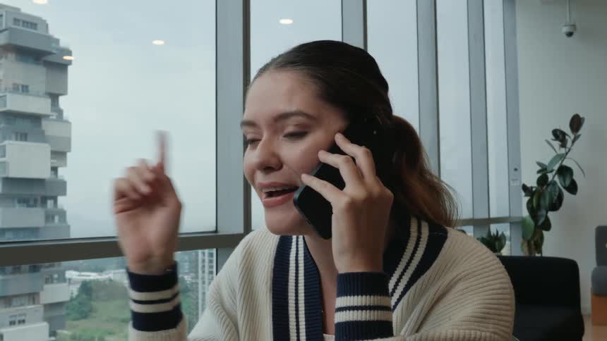 An excited young woman smiles and gestures while talking on her smartphone in a modern office with a cityscape view