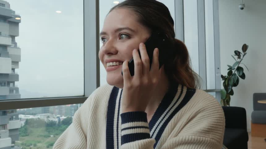 A smiling young woman in a modern office happily answers a phone call while looking out a large window