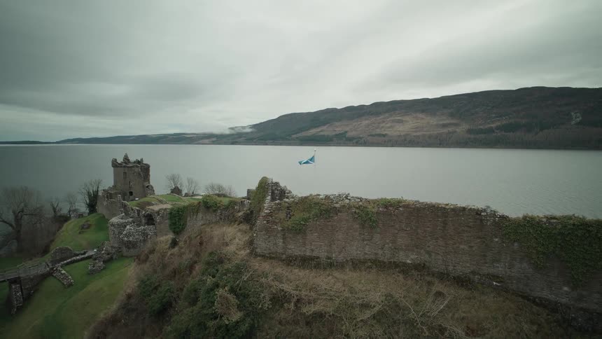 A dramatic aerial shot pulls back from the Scottish flag waving over the historic Urquhart Castle ruins on the shores of Loch Ness