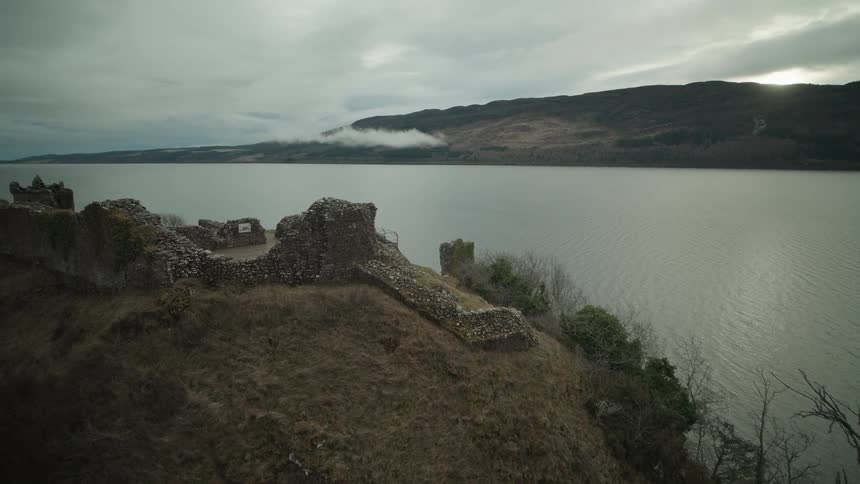 An aerial view slowly reveals the ancient ruins of Urquhart Castle on the moody banks of Loch Ness in Scotland