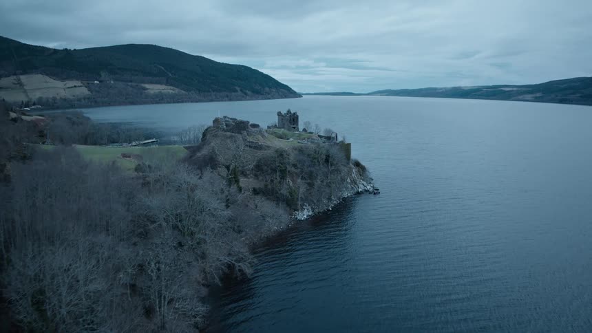 An aerial view flies over the dark waters of Loch Ness towards the historic Urquhart Castle ruins on a moody, overcast day in the Scottish Highlands