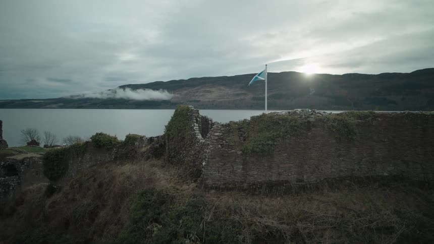 A dramatic aerial shot rises over the ancient stone walls of Urquhart Castle to reveal the Scottish flag flying over Loch Ness