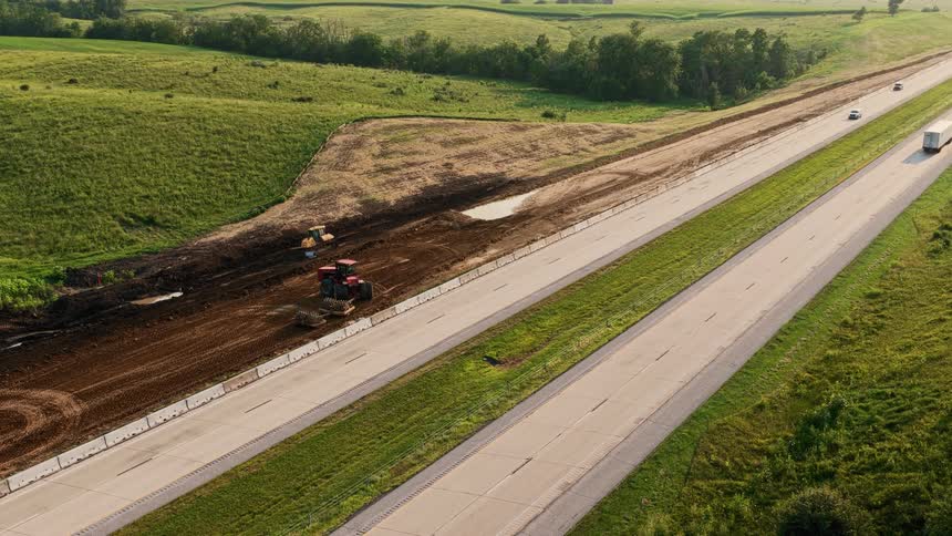 An aerial view shows heavy machinery working on a construction project alongside a busy highway in a rural landscape