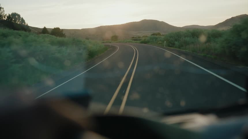 A scenic point-of-view drive along a winding country road through a grassy, hilly landscape during a beautiful sunset