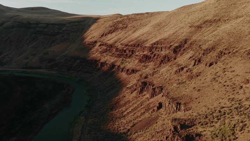 A static aerial shot captures a river snaking through an arid canyon as a long shadow creeps across the sunlit, rocky hillside