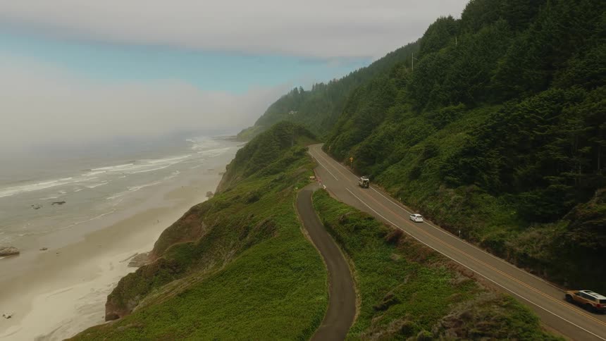 An RV and other vehicles drive along a scenic coastal highway carved into lush green cliffs overlooking the foggy ocean