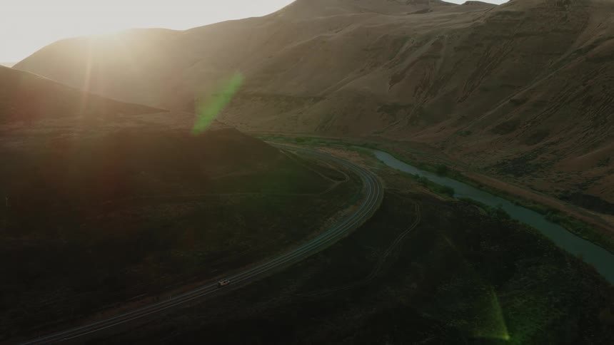 An aerial view of a lone car driving on a winding road through a mountain valley alongside a river as the sun sets