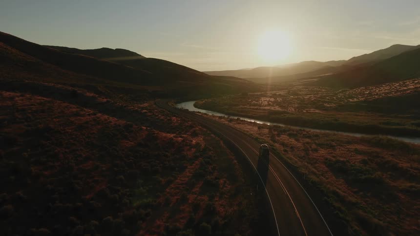 An aerial view shows a lone car driving on a scenic, winding road through a mountain valley alongside a river during a beautiful sunset