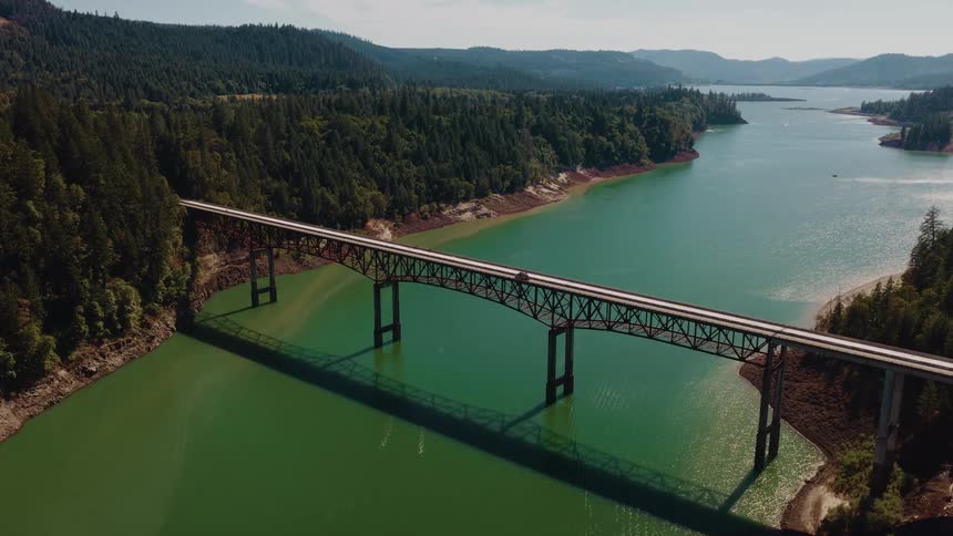 A scenic aerial shot captures a car driving over a steel truss bridge that crosses a wide green river surrounded by dense evergreen forests