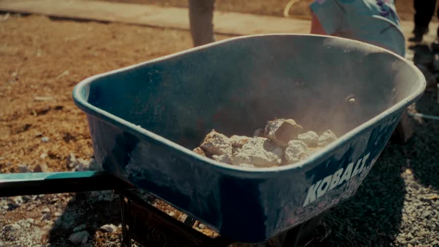 A person tosses rocks into a blue wheelbarrow in slow motion at a sunny work site
