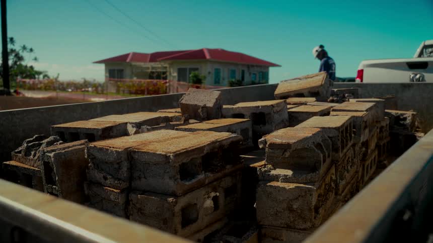 A close-up of weathered cinder blocks piled in a truck bed on a sunny day with a construction worker in the background