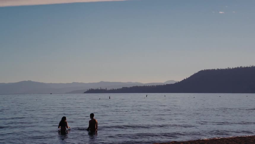 A couple in silhouette wades in the calm waters of a lake at dusk with mountains in the background