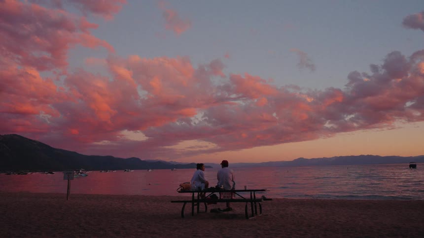 A serene wide shot shows a couple sitting on a picnic table on a sandy beach, enjoying a spectacular pink sunset over a calm lake