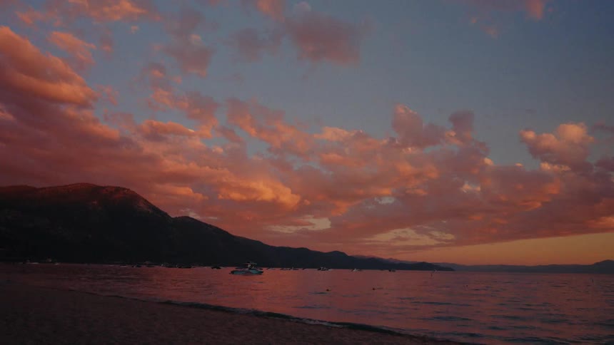A vibrant pink and orange sunset fills the sky with clouds over a calm mountain lake with boats anchored in the bay