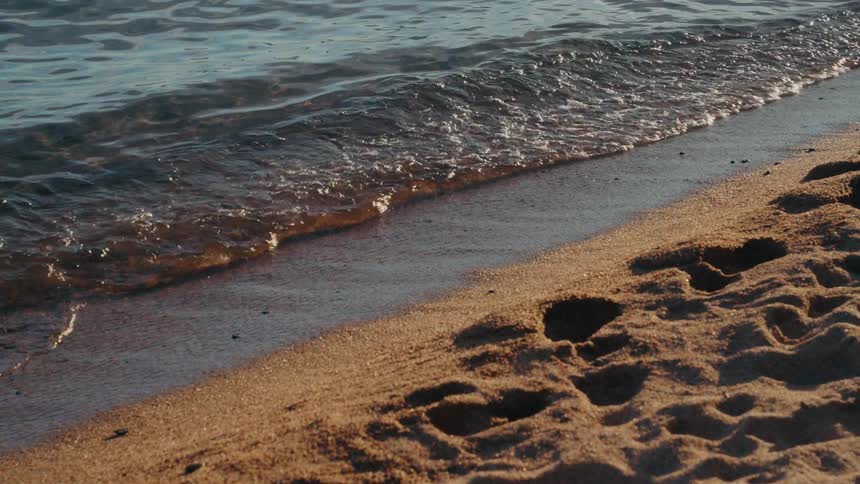 Peaceful slow motion footage captures gentle waves kissing a sandy shore dotted with footprints