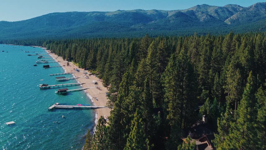 Stunning aerial view of the Lake Tahoe coastline with clear turquoise water, sandy beaches, private docks, and a dense pine forest