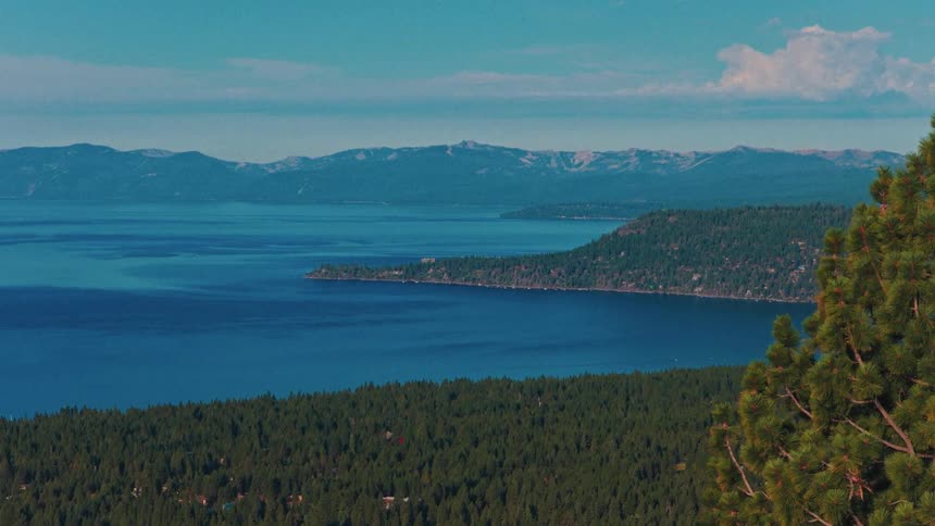 A majestic, slow-zoom view of the deep blue waters of Lake Tahoe, surrounded by a dense evergreen forest and mountain ranges under a clear sky