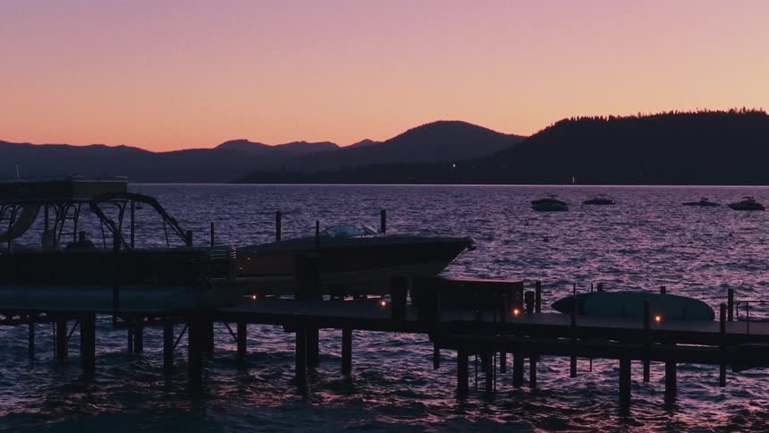 A slow zoom towards a pier with boats docked on a rippling lake, silhouetted against a magnificent purple and pink sunset sky