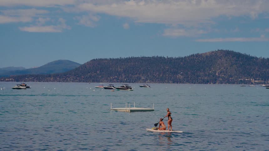 Two women in bikinis enjoy a leisurely day paddleboarding across a serene mountain lake surrounded by boats