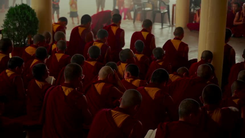 Buddhist monks in traditional red and yellow robes gather for prayer and study inside a dimly lit monastery