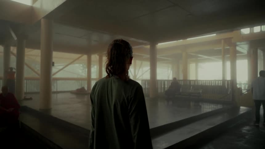 A lone figure stands in a misty, open-air temple, observing monks and devotees in a serene atmosphere