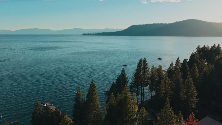 A slow aerial ascent over pine trees reveals the breathtaking expanse of a mountain lake at golden hour