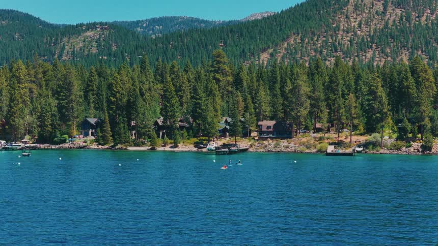 A serene view of a pristine blue mountain lake with people paddleboarding and boating near a shoreline dotted with pine trees and cabins
