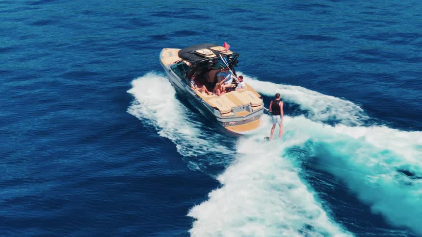 A thrilling aerial slow-motion shot captures a man wakeboarding behind a boat full of friends on the open sea before falling into the water