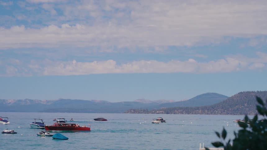 A vintage wooden boat and various other watercrafts float on the calm waters of Lake Tahoe, with pine-covered mountains in the background