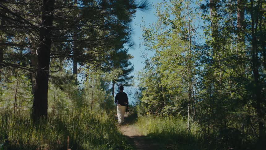 A man with a camera walks down a lush, sunlit forest path on an outdoor adventure