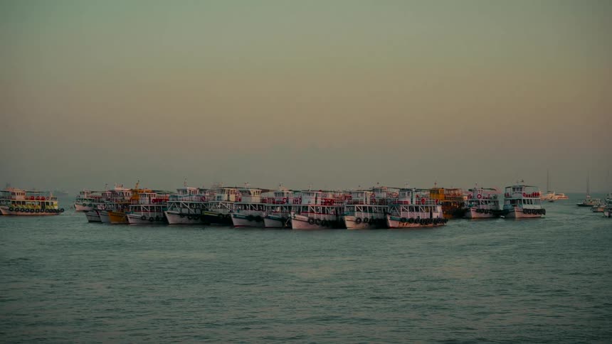 A large cluster of passenger ferries are moored together, bobbing on the calm sea during a hazy sunset