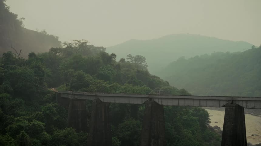 A tranquil, static shot of a railway bridge crossing a lush green valley amidst misty mountains