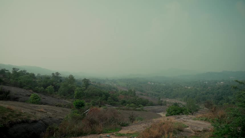 A static wide shot captures a hazy mountain valley landscape with rocky terrain and green forests