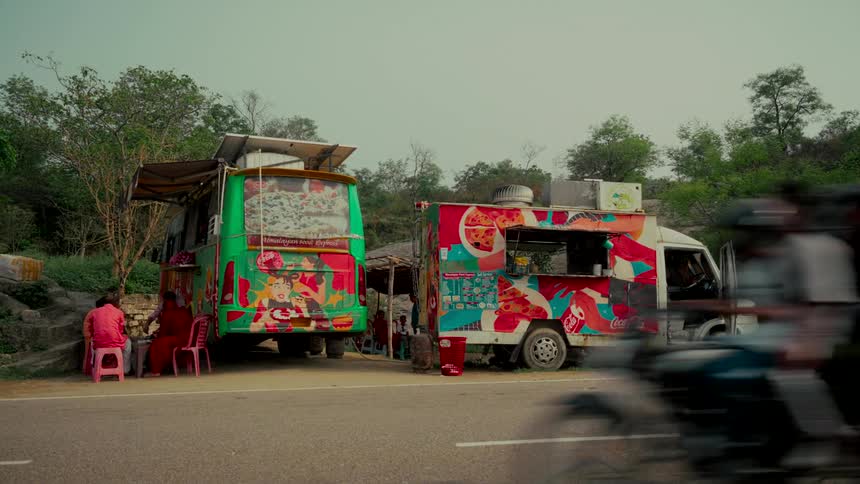 Two colorful food trucks are parked on a rural roadside in India as people sit and relax and traffic passes by