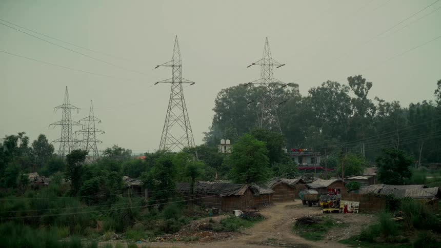 A rustic village with makeshift huts sits in a lush green landscape beneath towering electricity pylons on a cloudy day