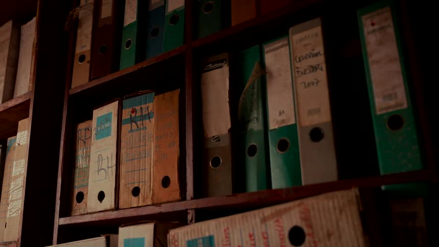 Old, dusty, and worn file binders and document boxes fill wooden shelves in a dark, mysterious archive room