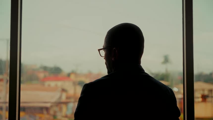 A businessman in silhouette stands thoughtfully, looking out a large window at the city view