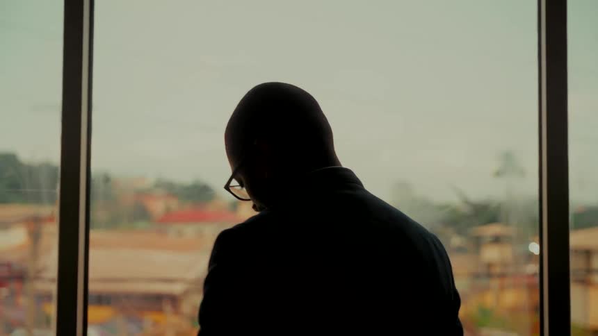 A silhouetted businessman in a suit makes a phone call from his office while looking out the window at the city below