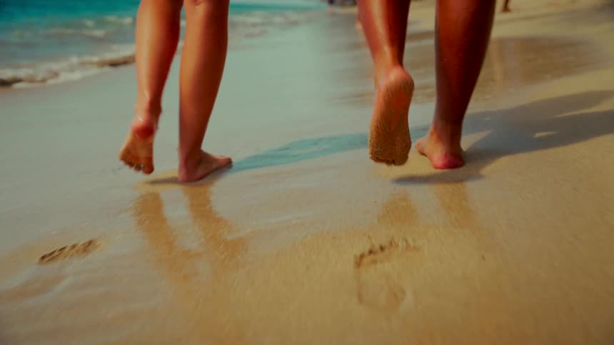 Slow motion close-up of two people's legs and feet as they walk barefoot on the wet sand at the ocean's edge