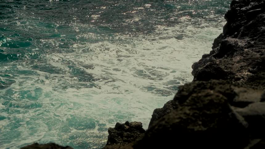 Sunlight glints off turbulent turquoise ocean waves crashing against a rocky shoreline in slow motion