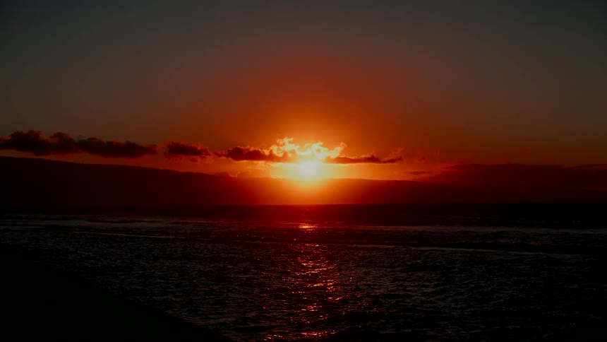 A vibrant orange sun sets over the ocean, casting a brilliant red reflection on the gentle waves