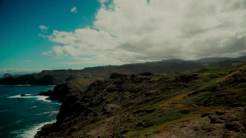 A slow pan reveals a rugged, green coastal landscape overlooking the vast blue ocean under a partly cloudy sky