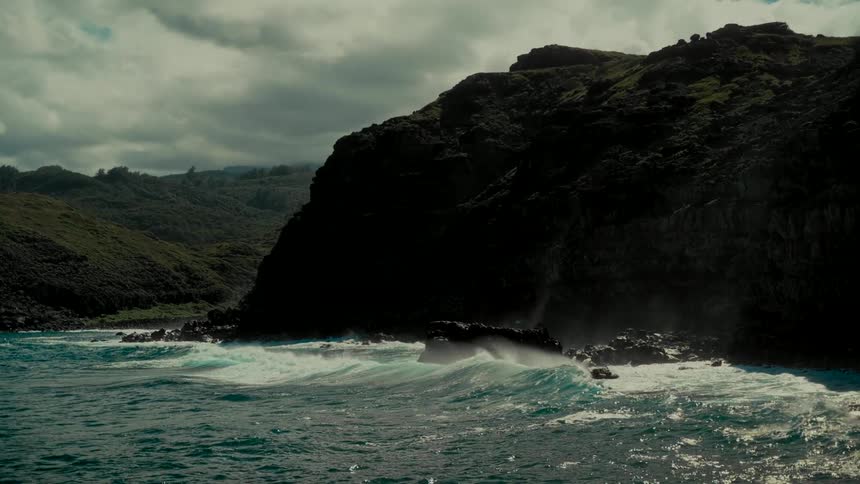 Dramatic slow-motion shot of powerful ocean waves crashing against a rugged, volcanic coastline under a cloudy sky
