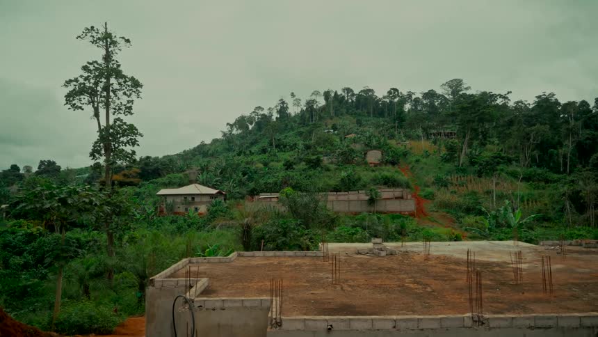 A wide shot shows a construction site with a concrete foundation overlooking a lush green, tree-covered hill in a rural village under an overcast sky