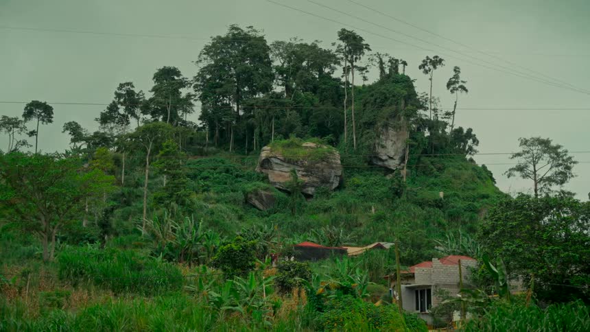 A static wide shot of a lush green tropical hillside with prominent rock formations overlooking a small settlement
