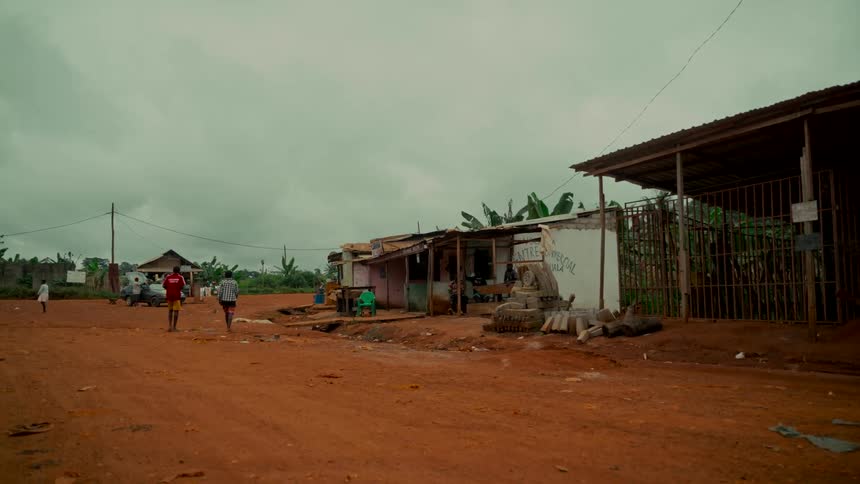 People walk down a red dirt road through a small, rural African village on a cloudy day