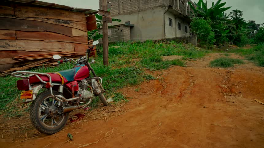 A still shot shows a red motorcycle parked on a dirt road next to a wooden shack in a rural village