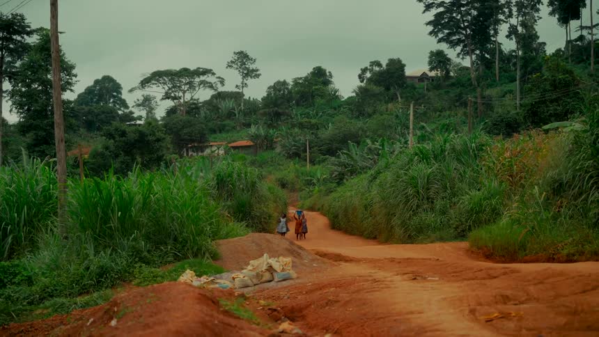 A wide shot shows a woman and two children walking away on a red dirt road through a lush, green rural village in Africa