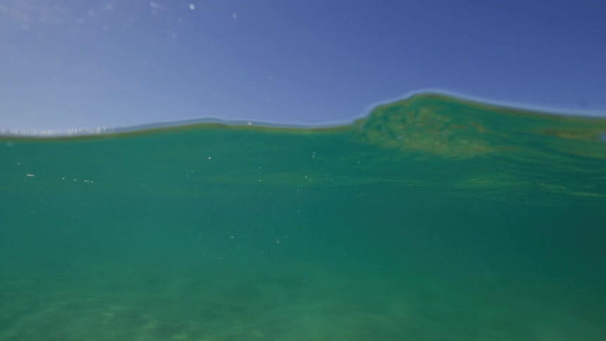 A serene slow-motion split-screen view of a gentle ocean wave rolling over the camera under a clear blue sky