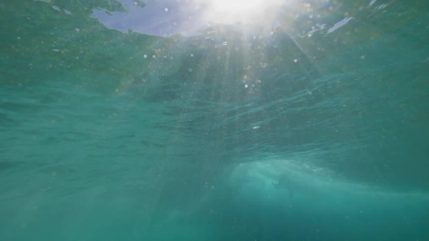An ethereal underwater perspective captures shimmering sunbeams piercing through the calm, turquoise ocean surface in slow motion